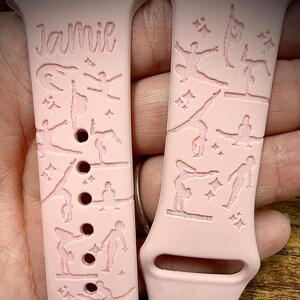 May include: A light pink silicone watch band featuring laser-engraved gymnastics poses and small star accents. The name Jamie is engraved at the top of the left strap. The design includes various gymnasts performing floor and beam routines.