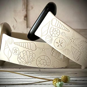 May include: A white silicone watch band featuring an embossed pattern of various seashells, starfish, and sand dollars. The band includes a black connector piece at the top and a metallic gold connector piece on the left side, showcasing a beach theme.