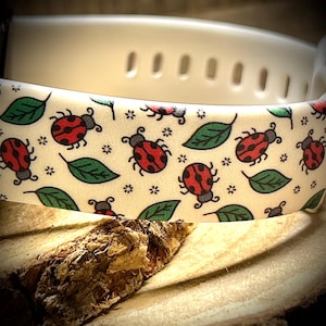 May include: A white watch band with a repeating pattern of red ladybugs, green leaves, and small black flowers. The band is made of a flexible material and has a series of holes for adjustment.