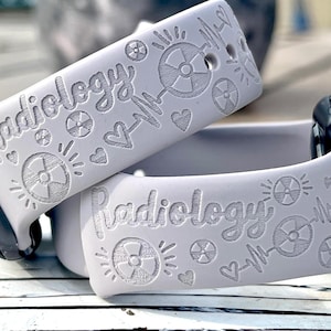 May include: A light gray silicone watch band featuring laser-engraved designs including the word Radiology in a cursive script, radiation warning symbols, small hearts, and EKG heart rhythm lines across the surface of the flexible material.
