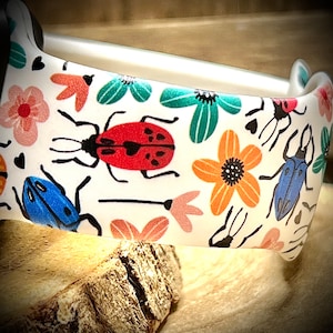 May include: White watch band with a colorful floral and bug pattern. The pattern includes red, blue, green, orange, and pink flowers and bugs.
