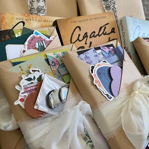 May include: A stack of wrapped books with white ribbon bows. The books are wrapped in brown paper and have various stickers and decorations. The books are titled "The Queen of Mystery" and feature the author Agatha Christie.