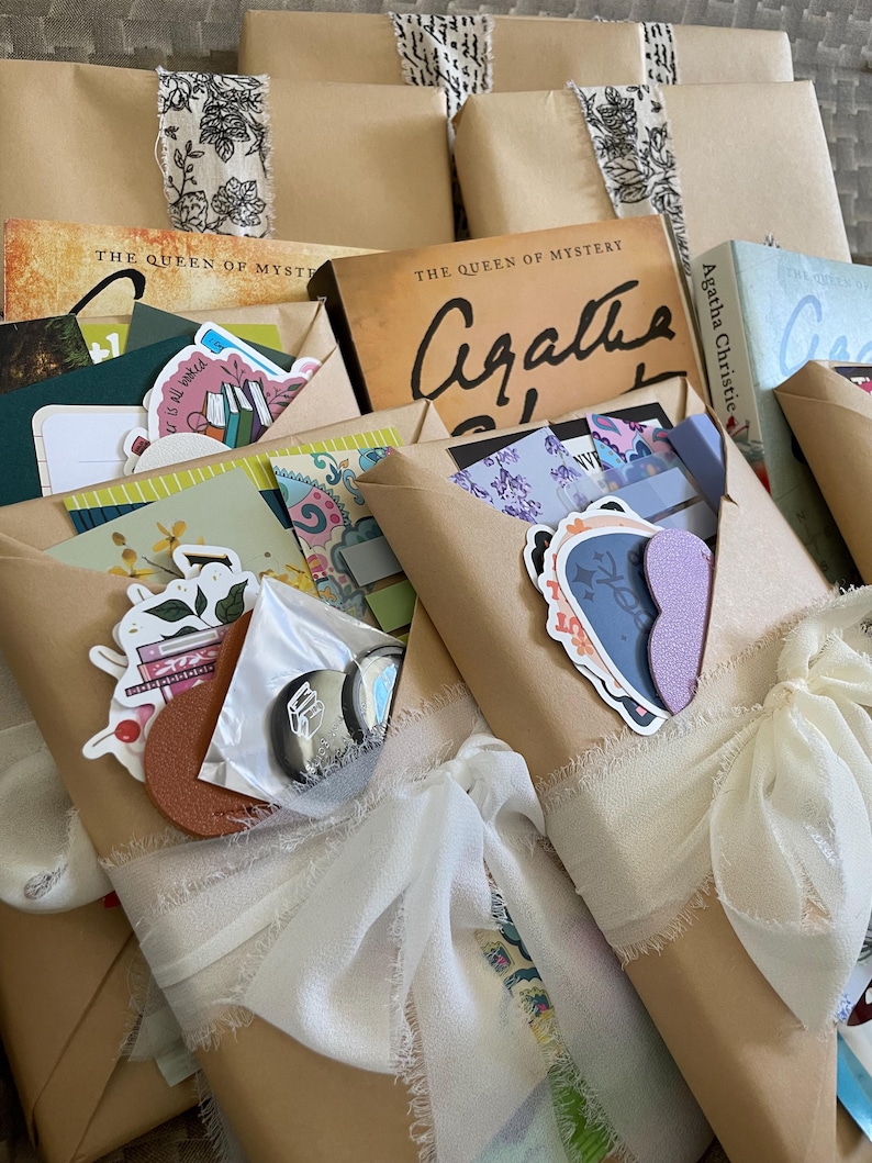 May include: A stack of wrapped books with white ribbon bows. The books are wrapped in brown paper and have various stickers and decorations. The books are titled "The Queen of Mystery" and feature the author Agatha Christie.