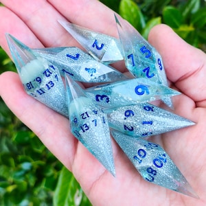 Op de afbeelding: Een set van zeven doorschijnende, langwerpige veelzijdige dobbelstenen met scherpe punten. Elke dobbelsteen heeft een behuizing van heldere hars gevuld met een lichtblauwe, bubbelachtige en glinsterende textuur. De cijfers zijn in donkerblauw gedrukt.
