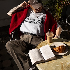 May include: A person wearing a white t-shirt with the text "HALTE DEINE UMWELT SAUBER" and a swastika symbol. The person is sitting at a table with a book, a croissant, and a glass of iced coffee.