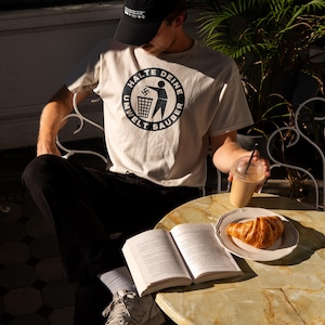 Może przedstawiać: Osoba w białej koszulce z czarnym kołem z napisem "Halte deine Umwelt sauber" i czarną czapką bejsbolową siedzi przy stole z książką, croissantem i szklanką lodowatej kawy.