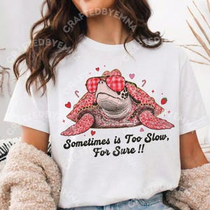 Peut inclure: T-shirt blanc avec une tortue de dessin animé portant des lunettes de soleil à carreaux rouges. La tortue est rose avec une carapace à motifs. Le t-shirt porte l'inscription "Sometimes is Too Slow, For Sure !!" avec des cœurs et des cannes en bonbon.