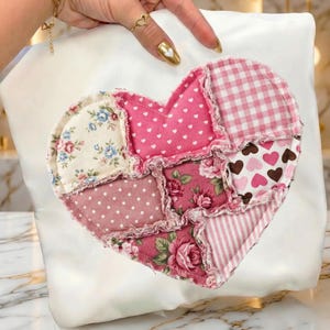 Peut inclure: Coussin blanc avec un cœur en patchwork. Le cœur est composé de différents tissus roses, crème et marron, avec des motifs floraux, à pois et en forme de cœur. Les bords du cœur sont effilochés, ajoutant une touche rustique.