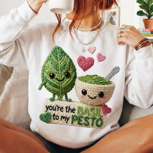 Peut inclure: Sweat-shirt blanc avec un motif crochet représentant une feuille verte souriante et un bol de pesto. Le texte "you're the basil to my pesto" est en dessous. Des cœurs roses au crochet sont également inclus.