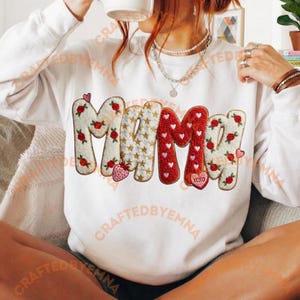 Peut inclure: Sweat-shirt blanc avec le mot "MAMA" dans un design décoratif. Les lettres sont ornées de roses, de cœurs et d'étoiles. Une fraise et un "XOXO" en forme de cœur complètent le design.