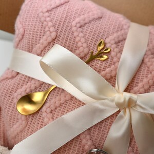 May include: A pink knitted blanket with a white satin ribbon tied around it. A gold spoon and a gold twig with leaves are on top of the blanket.
