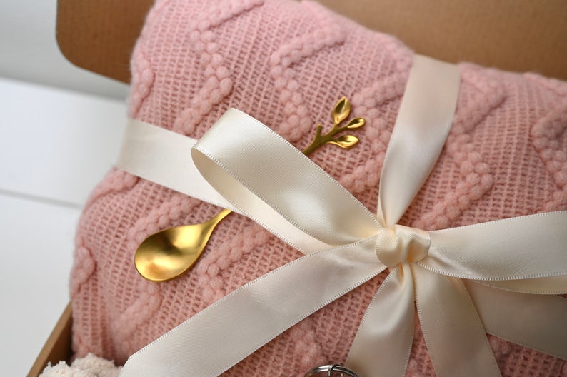 May include: A pink knitted blanket with a white satin ribbon tied around it. A gold spoon and a gold twig with leaves are on top of the blanket.