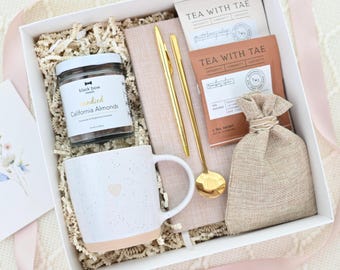 Puede incluir: Una caja de regalo blanca llena de una taza, un tarro de almendras confitadas, sobres de té, una pequeña bolsa de arpillera, un cuaderno y bolígrafos y cuchara dorados. La taza tiene un diseño de corazón y una base rosa claro. La caja está decorada con una cinta rosa.