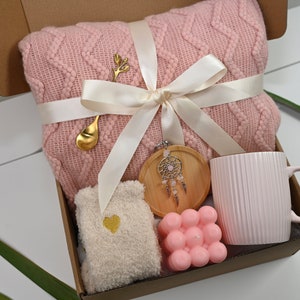 May include: A pink knitted throw blanket, a pink mug, a wooden coaster with a dreamcatcher, a pink bath bomb, and a pair of white socks with a gold heart in a brown gift box.