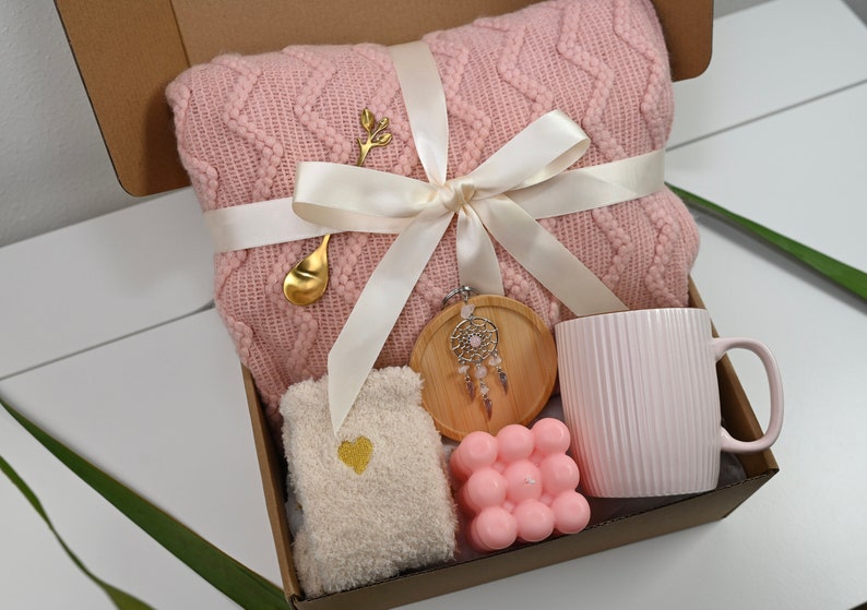 May include: A pink knitted throw blanket, a pink mug, a wooden coaster with a dreamcatcher, a pink bath bomb, and a pair of white socks with a gold heart in a brown gift box.