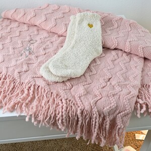 May include: A pink knitted throw blanket with a fringe border and a white fuzzy sock with a gold heart on it.