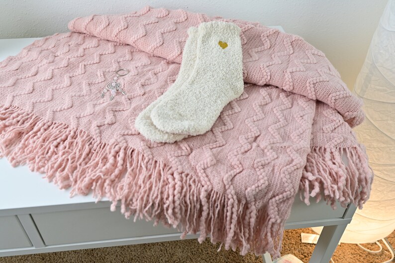 May include: A pink knitted throw blanket with a fringe border and a white fuzzy sock with a gold heart on it.