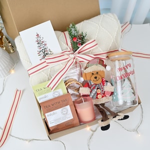 May include: A Christmas gift box with a white blanket, tea packets, a candle, a glass with "Merry Christmas" text, and a teddy bear ornament. The box is tied with a red and white striped ribbon and includes a card with a Christmas tree illustration.
