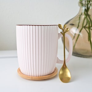 May include: A pink ceramic mug with a ribbed design and a brown rim. The mug is on a wooden coaster and has a gold spoon with a leaf design.