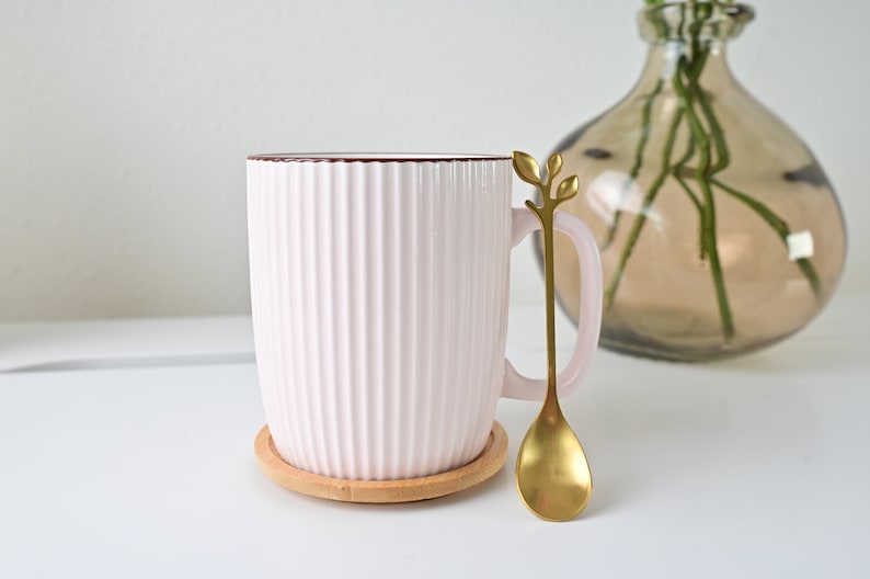 May include: A pink ceramic mug with a ribbed design and a brown rim. The mug is on a wooden coaster and has a gold spoon with a leaf design.