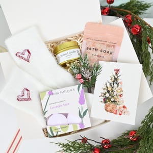 Könnte beinhalten: Eine Geschenkbox mit Pflegeprodukten. Sie enthält weiße Socken mit gestickten Herzen, eine goldene Kerze, Badesalz, Dusch-Trockenshampoo und eine Weihnachtskarte. Die Box ist mit Grün und roten Kugeln verziert.