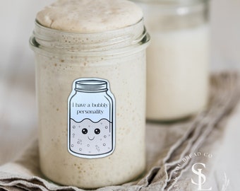 May include: Two glass jars filled with a bubbly white liquid. The jar in the foreground has a sticker on it that says "I have a bubbly personality" and depicts a cartoon jar with a smiling face. The jars are sitting on a light brown linen cloth.