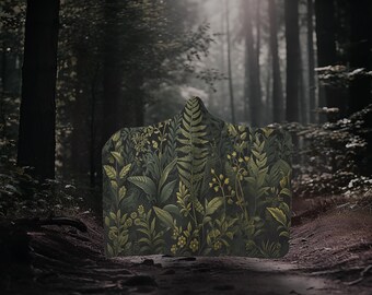 May include: A dark green hooded blanket with a botanical print featuring ferns and other plants in shades of green and yellow. The blanket is lying on a path in a forest.