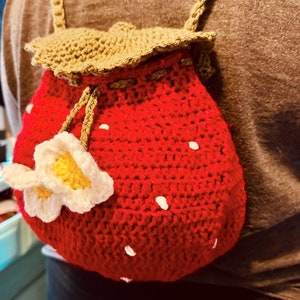 Könnte beinhalten: Eine rote gehäkelte Tasche in Form einer Erdbeere mit weißen Punkten und einem grünen gehäkelten Blatt oben. Die Tasche hat eine weiße gehäkelte Blume mit gelber Mitte an der Seite.
