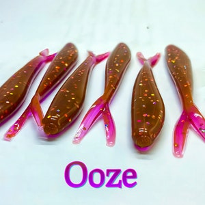 May include: Five brown and pink fishing lures with glitter. The lures are shaped like fish and have a pink tail. The text "Ooze" is written in pink letters below the lures.