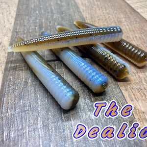 May include: A close-up of five brown and white soft plastic fishing lures with blue glitter. The lures are arranged in a row on a wooden surface. The text "The Dealio" is visible in the lower right corner of the image.