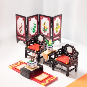 Puede incluir: Conjunto de muebles en miniatura en negro y rojo, hechos con pequeñas cuentas de colores. El conjunto incluye sillas con cojines rojos, una pantalla con diseños florales y una pequeña mesa. Pequeñas plantas en macetas decoran el conjunto.
