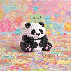 May include: A handmade panda bear figurine made of small, colorful beads. The panda is black and white with pink accents and is sitting down. The background is a colorful, patterned surface with cartoon characters.