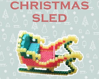 以下が含まれることがあります： 小さなプラスチックビーズを融合させて作られた、カラフルなクリスマスのそり。そりは赤、黄、緑、青で、上に赤い文字で「CHRISTMAS SLED」と書かれています。背景は、クリスマスをテーマにしたイラストが描かれた水色です。
