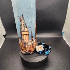 Puede incluir: Un vaso cilíndrico alto con un fondo azul claro y una imagen detallada de un castillo. Se combina con una pequeña escultura azul y negra que sostiene un libro en miniatura. El vaso y la escultura están sobre una plataforma negra.