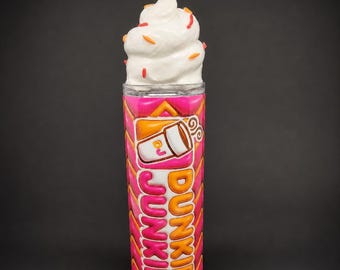 Könnte beinhalten: Ein zylindrischer Becher mit einem pinkfarbenen und orangefarbenen Zickzackmuster und dem Text "DUNKIE JUNKIE" in orangefarbenen und pinkfarbenen Buchstaben. Der Becher zeigt eine Kaffeetasse und hat einen Deckel in Form von weißer Schlagsahne mit orangefarbenen Streuseln.