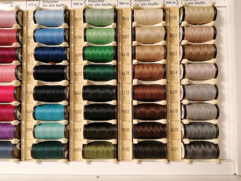May include: A close-up of a variety of spools of thread in different colors, including pink, red, blue, green, brown, and gray. The spools are arranged in rows and columns, and each spool has a label with a number on it. The labels also indicate that the thread is polyester and suitable for all fabrics. The spools are 100 meters long.