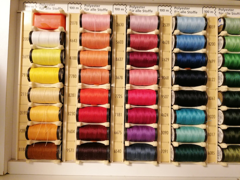 May include: A close-up of a box of 100-meter spools of polyester thread in various colors. The spools are arranged in rows and columns, with each spool labeled with a number.