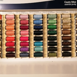 May include: A close-up of a box of Coats Mez thread spools. The spools are arranged in rows and columns, and each spool is labeled with a number and the color of the thread. The thread is made of polyester and is 100 meters long. The box is labeled "Coats Mez" and "nat&uuml;rlich farbig".