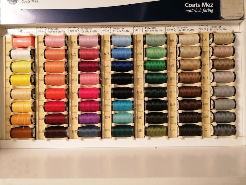 May include: A close-up of a box of Coats Mez thread spools. The spools are arranged in rows and columns, and each spool is labeled with a number and the color of the thread. The thread is made of polyester and is 100 meters long. The box is labeled "Coats Mez" and "nat&uuml;rlich farbig".