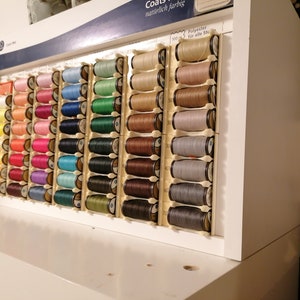 May include: A close-up of a wall-mounted display of colorful spools of thread. The spools are arranged in rows and columns, and each spool is labeled with a number. The text on the display reads "Coats" and "manufaktur farbig".