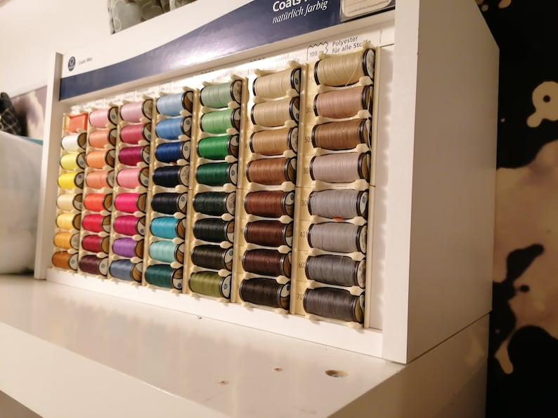 May include: A close-up of a wall-mounted display of colorful spools of thread. The spools are arranged in rows and columns, and each spool is labeled with a number. The text on the display reads "Coats" and "manufaktur farbig".