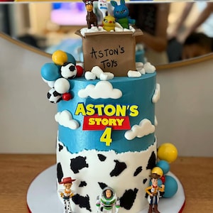 May include: A birthday cake with blue and white frosting, decorated with toy figures and the text "ASTON'S STORY 4". The cake has a cow-print design on the bottom tier and a brown box on top that reads "Aston's Toys".