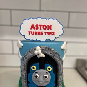 May include: A blue and gray Thomas the Tank Engine cake with a white cloud topper that says "Aston Turns Two!" in red letters.