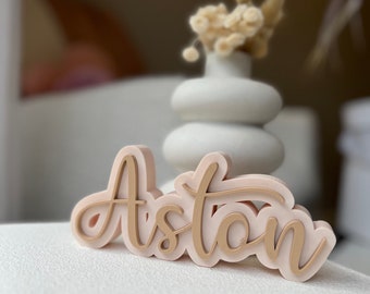 May include: A light pink wooden name sign with the name "Aston" written in a cursive font in a light brown color.