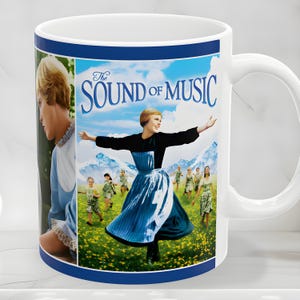 May include: A white ceramic mug featuring a blue border and a printed graphic from The Sound of Music. The image displays a person with short hair in a black and blue dress standing in a field with children, set against a backdrop of mountains.