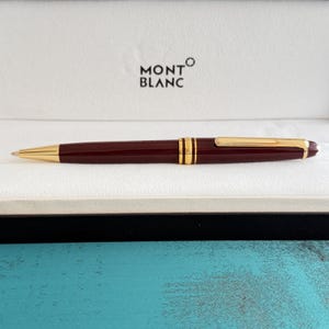 Puede incluir: Un bolígrafo de color burdeos con detalles en tono dorado, que incluyen un clip y una banda central. El bolígrafo está colocado horizontalmente sobre un fondo blanco con el logotipo de Montblanc en negro, sobre una superficie turquesa texturizada.