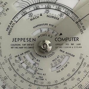 May include: A Jeppesen Model R.2 Computer for calculating true airspeed, density altitude, and true altitude. The circular slide rule has a white face with black markings and a silver metal center. The computer is labeled "Jeppesen Model R.2 Computer" and "Copyright 1955 Ray Lahr".