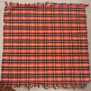 Puede incluir: Una manta a cuadros rojos, amarillos y negros con un borde con flecos. La manta presenta un patrón de tartán clásico, con gruesas líneas negras que se cruzan con líneas amarillas más finas sobre un fondo rojo. El borde con flecos añade un toque decorativo.