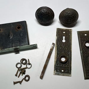 Może przedstawiać: Asortyment zabytkowych okuć do drzwi, w tym dwa ozdobne gałki do drzwi, mechanizm zamka, dwie dekoracyjne płytki, drążek zatrzaskowy oraz różne śruby i podkładki. Okucia są metalowe i noszą ślady wieku i zużycia.