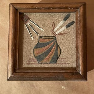 May include: A framed artwork featuring a stylized pot with black, orange, and blue stripes. Matches and a bundle of sticks are depicted above the pot. The frame is made of dark wood.